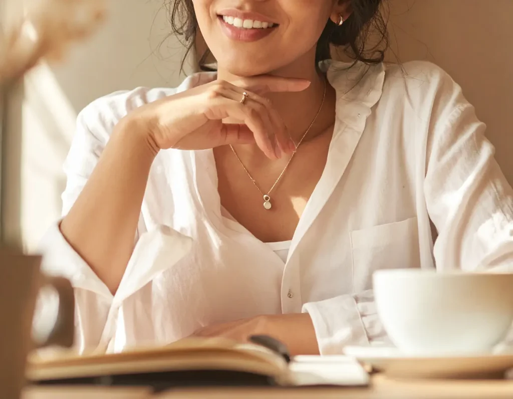 Smiling woman in a white blouse wearing a delicate necklace and simple ring while enjoying coffee at a café, representing casual everyday jewelry. Design One Jewelers, Clearwater FL.
