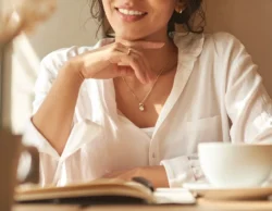 Smiling woman in a white blouse wearing a delicate necklace and simple ring while enjoying coffee at a café, representing casual everyday jewelry. Design One Jewelers, Clearwater FL.