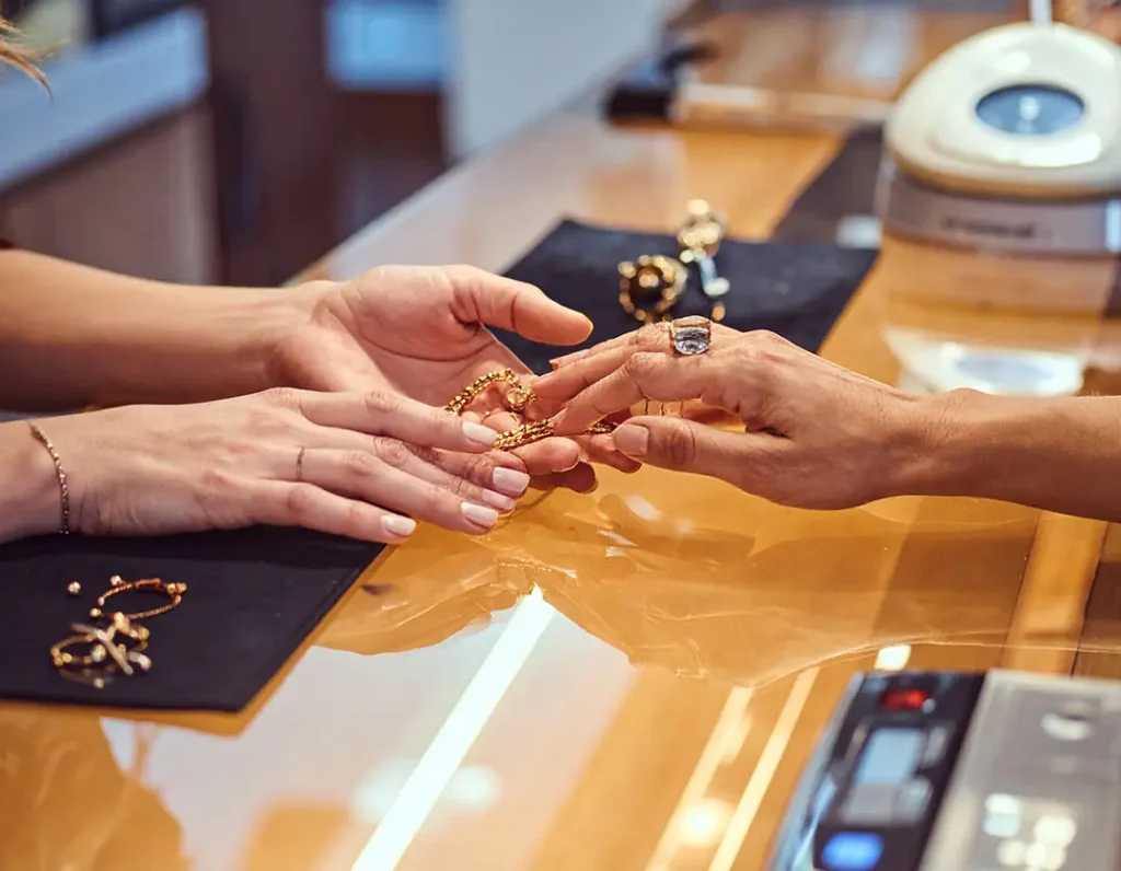Jeweler and customer’s hands exchanging gold jewelry across a warm glass counter.