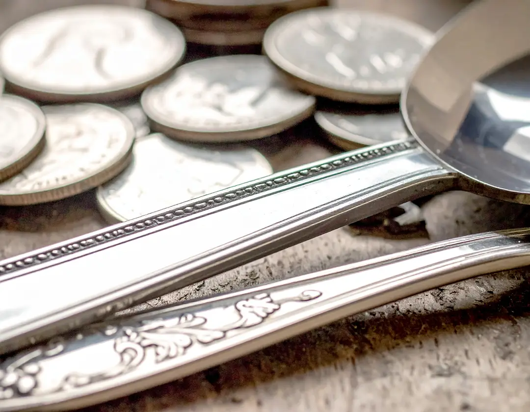 Macro shot of sterling silver flatware with engraved detailing, coins softly blurred in the background.
